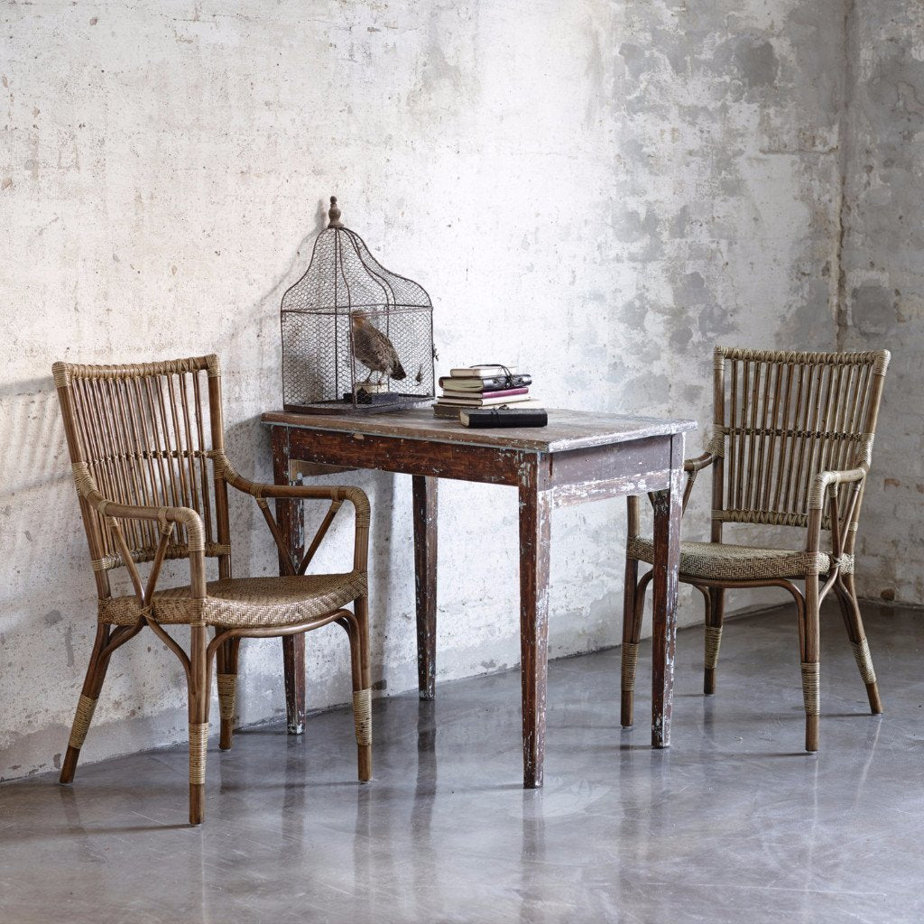 Rustic wooden table with weathered finish, paired with woven rattan chairs against a textured concrete backdrop.
