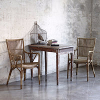 Rustic wooden table with weathered finish, paired with woven rattan chairs against a textured concrete backdrop.