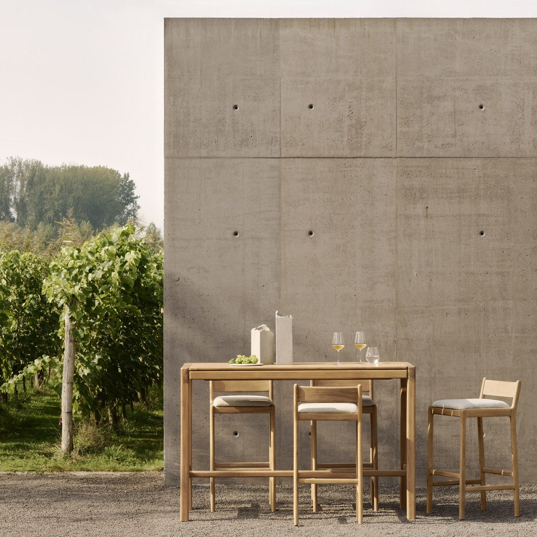 "Natural wood bar set with neutral cushions against a textured concrete wall, surrounded by greenery."