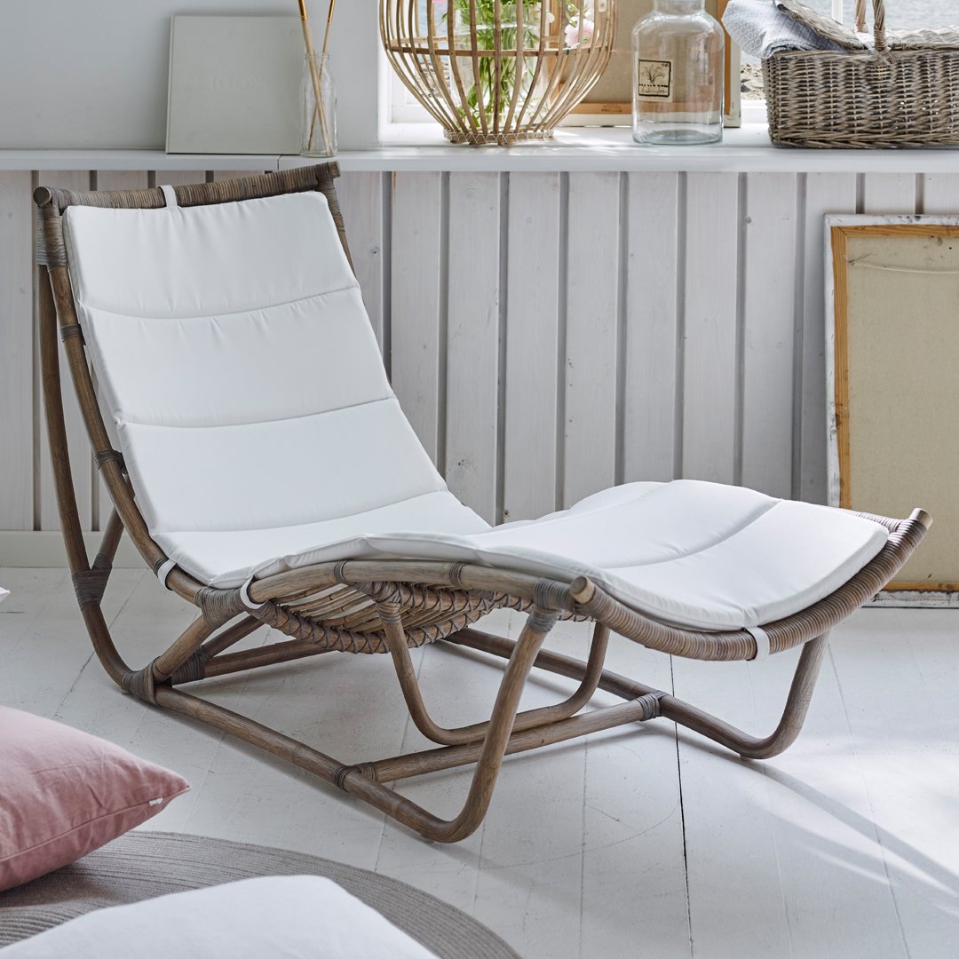 White cushioned lounge chair with a natural wood frame, set in a serene, minimalist interior with soft pastel accents.
