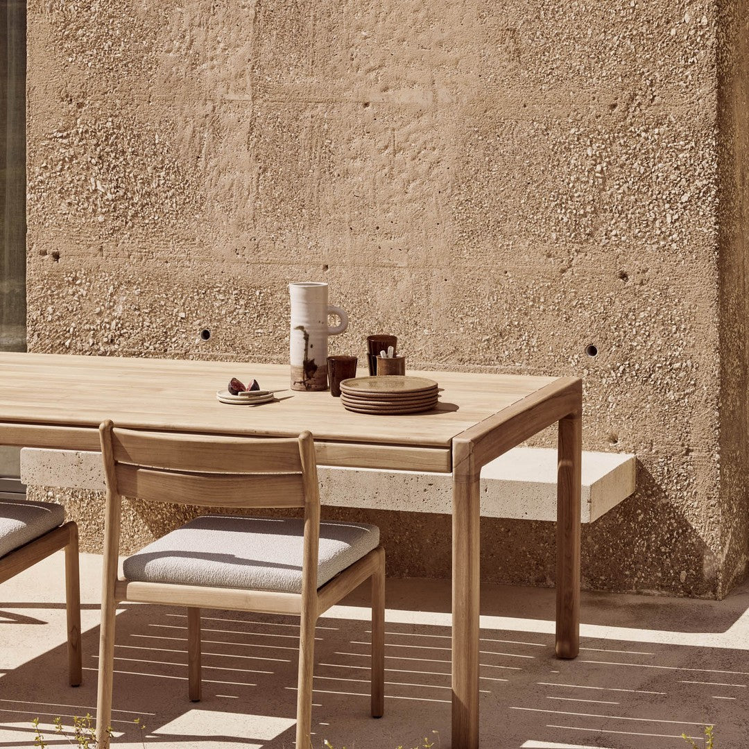 Natural wood table and chair with gray cushions, featuring earth-toned plates and a textured beige wall in background.
