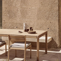 Natural wood table and chair with gray cushions, featuring earth-toned plates and a textured beige wall in background.