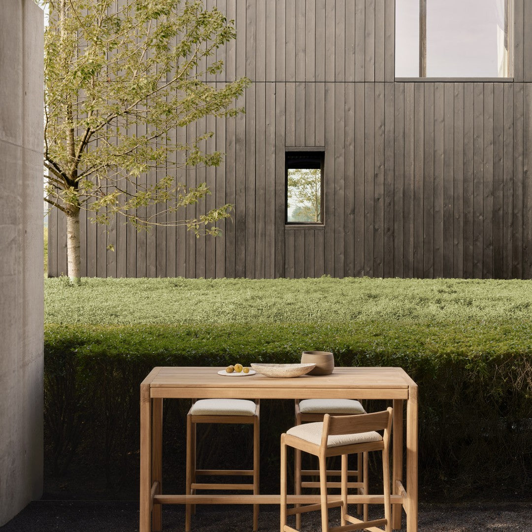 Natural wood table and chairs in neutral tones, set against a modern grey wooden backdrop and lush green grass.