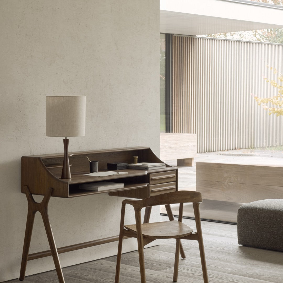 Mid-century modern desk in rich brown wood, paired with a soft beige lamp, set against a textured light gray wall.