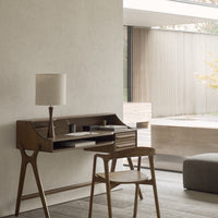 Mid-century modern desk in rich brown wood, paired with a soft beige lamp, set against a textured light gray wall.