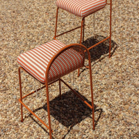 Striped upholstery in red and white on modern orange metal bar stools, set against a gravel background.