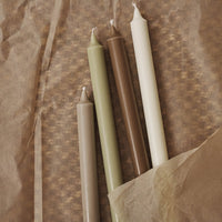 "Set of four tapered candles in earthy tones: olive green, brown, cream, and white on textured brown paper."