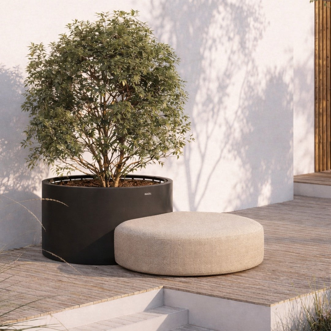 Beige round ottoman next to a black planter, set against a light textured wall and natural greenery.