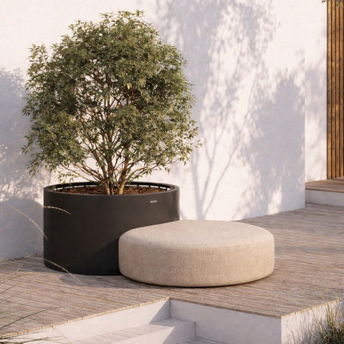 Beige round ottoman next to a black planter, set against a light textured wall and natural greenery.