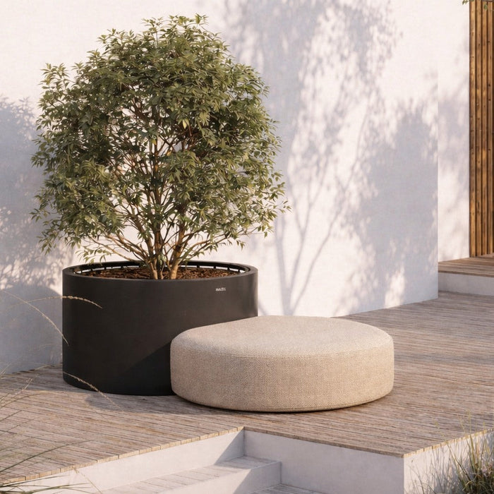 Beige round ottoman next to a black planter, set against a light textured wall and natural greenery.
