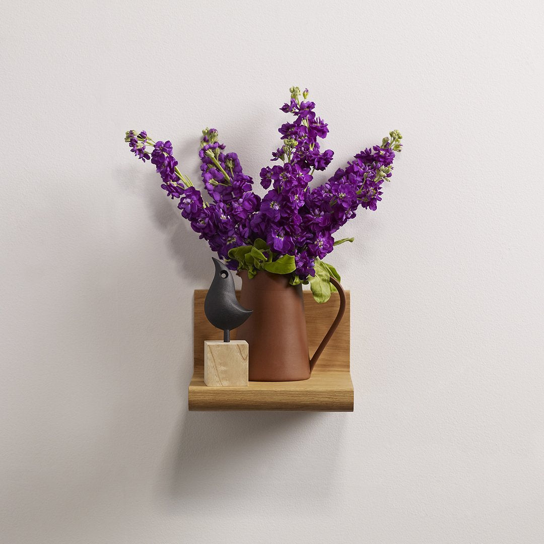 Purple flowers in a brown ceramic pitcher on a wooden shelf, set against a light background.