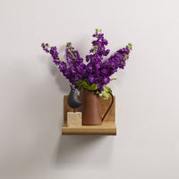 Purple flowers in a brown ceramic pitcher on a wooden shelf, set against a light background.