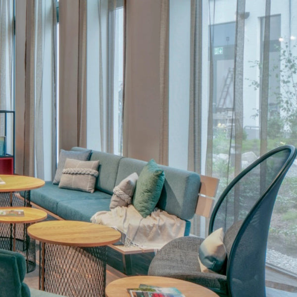 "Modern seating area with teal upholstery, colorful cushions, and light wood tables against soft, neutral curtains."