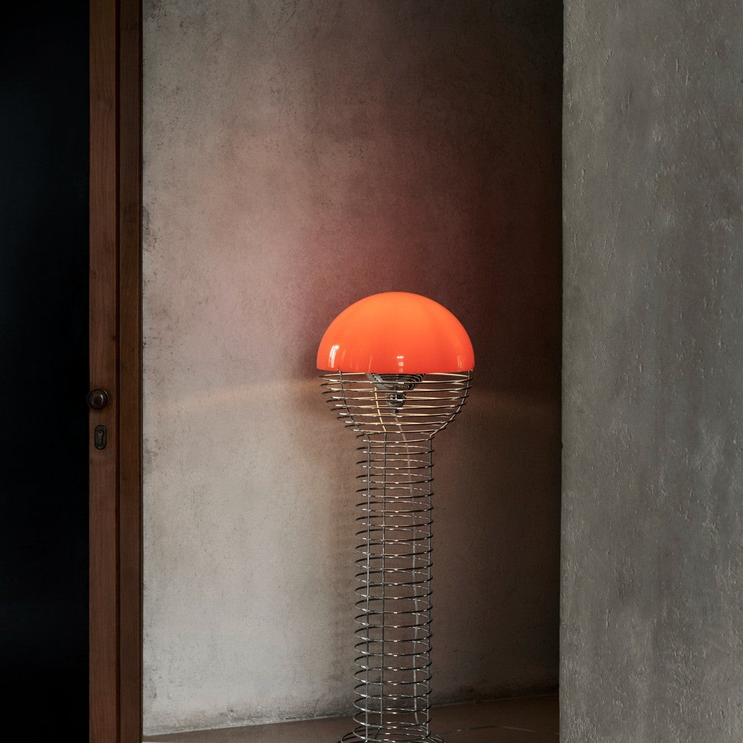 Modern lamp with a vibrant orange dome shade and sleek silver wire stand against a textured gray wall.