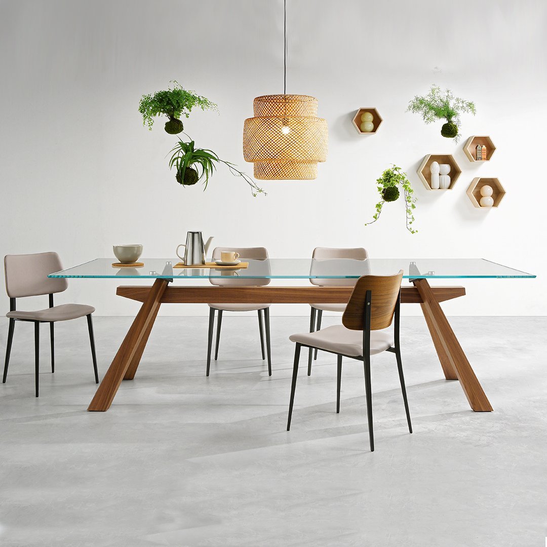 Minimalist glass dining table with wooden legs, complemented by light beige chairs and natural greenery accents.