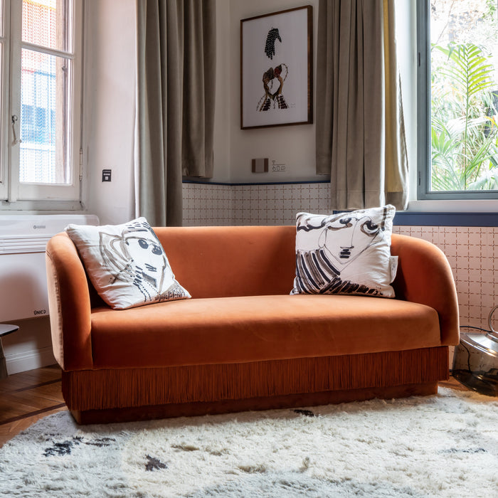 Rust-orange velvet sofa with fringe detailing and patterned black and white throw pillows. Soft, textured rug below.
