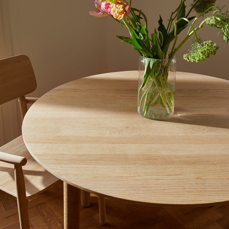 Natural light wood round table with smooth, light grain pattern, paired with a simple chair and green floral arrangement.