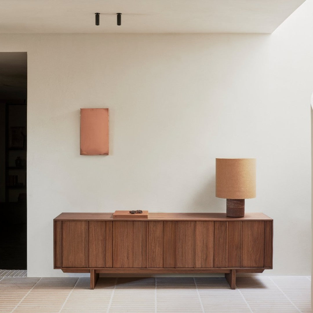 Wooden sideboard with warm brown tones, complemented by a beige lamp and copper wall art on a neutral background.