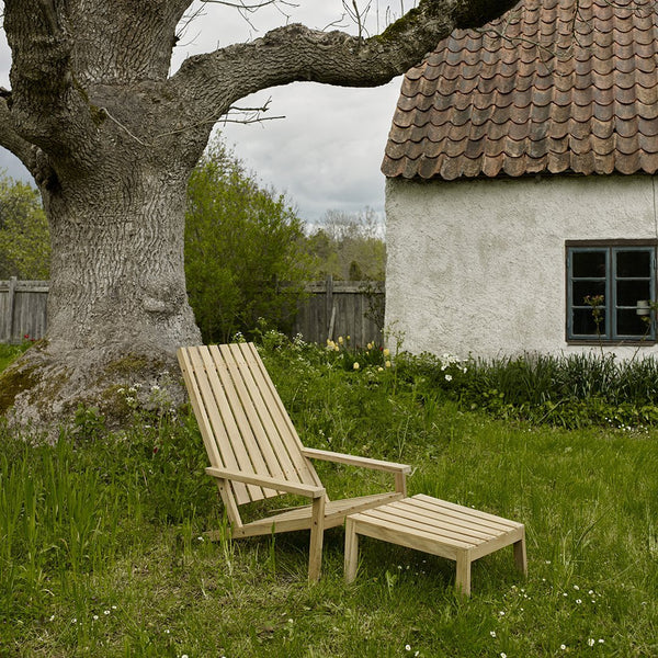 Natural wood lounge chair and ottoman in light beige, set against a green grass backdrop and textured brown roof.