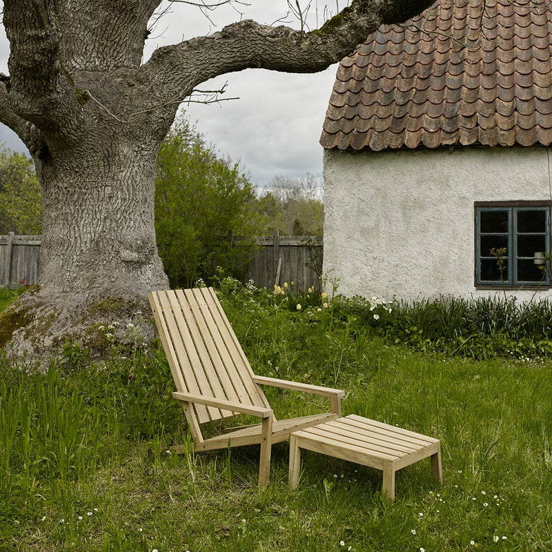 Natural wood lounge chair and ottoman in light beige, set against a green grass backdrop and textured brown roof.