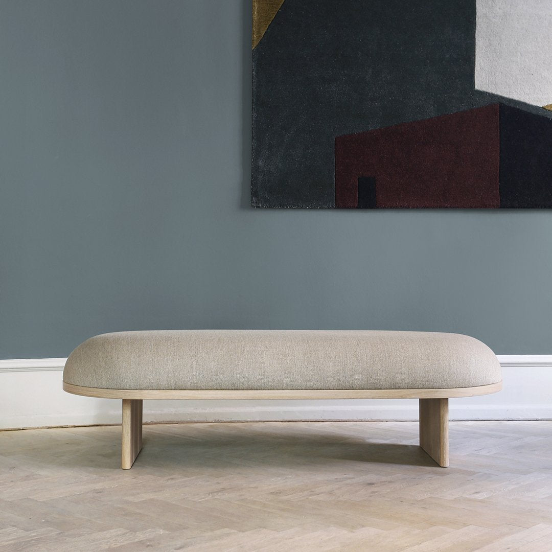 Light beige minimalist bench with smooth curves against a muted gray wall and abstract art featuring rich colors.