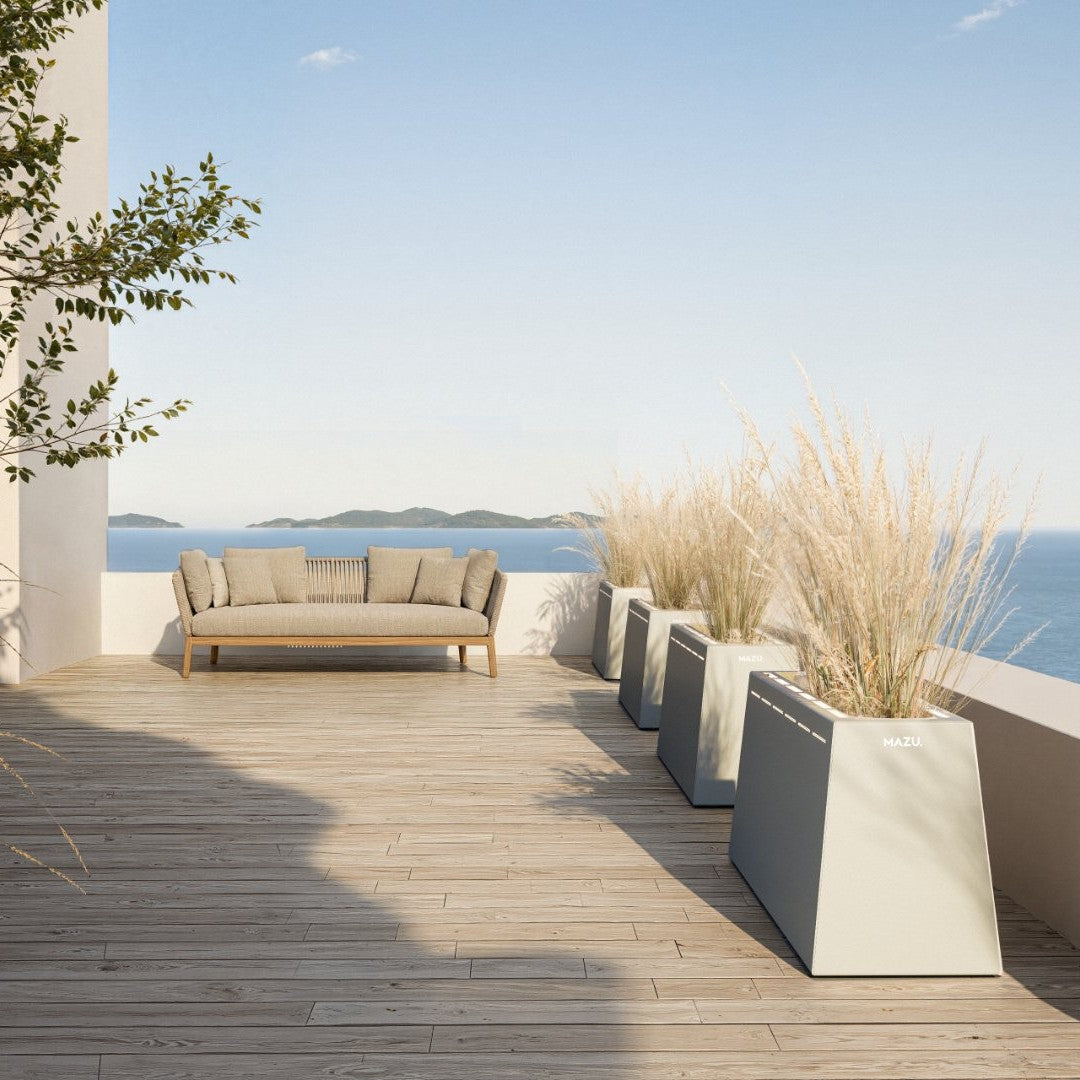 Beige outdoor sofa with light gray planters and natural wood decking, accented by green plants against a blue sky.
