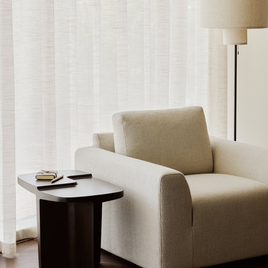 Beige textured armchair with a dark wooden side table, against sheer light curtains. Modern and minimalist aesthetic.