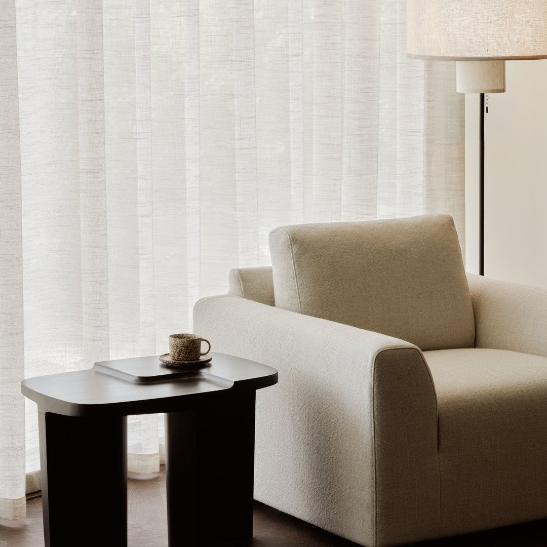 Beige textured armchair paired with a dark wood side table and sheer white striped curtains, creating a warm, cozy atmosphere.
