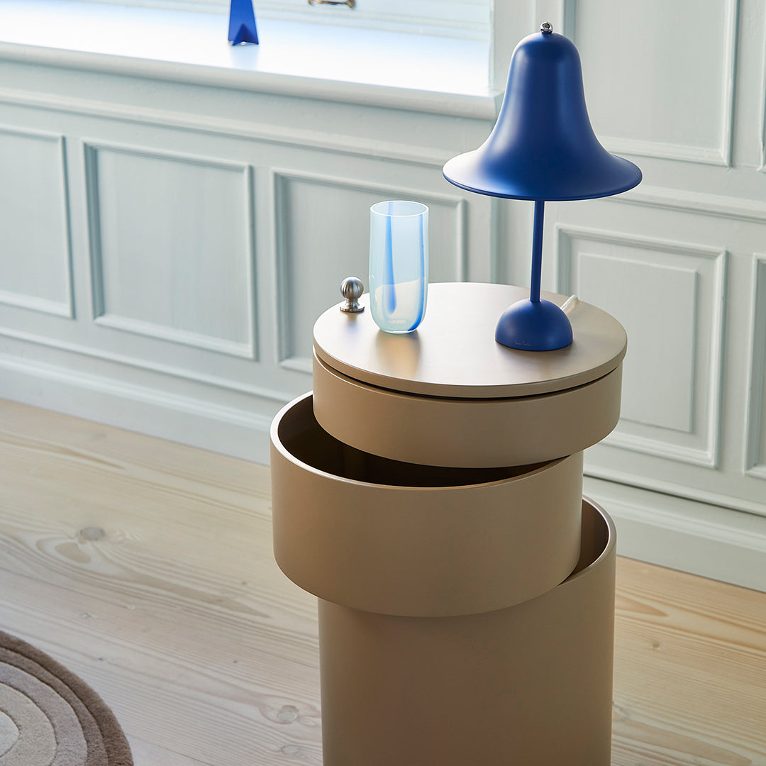 Beige cylindrical side table with blue lamp and glass vase, featuring smooth surfaces and modern, minimalist design.