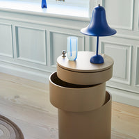 Beige cylindrical side table with blue lamp and glass vase, featuring smooth surfaces and modern, minimalist design.