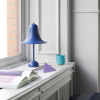 Blue bell lamp, teal cup, purple triangle, and patterned book on a light gray windowsill against a soft gray backdrop.