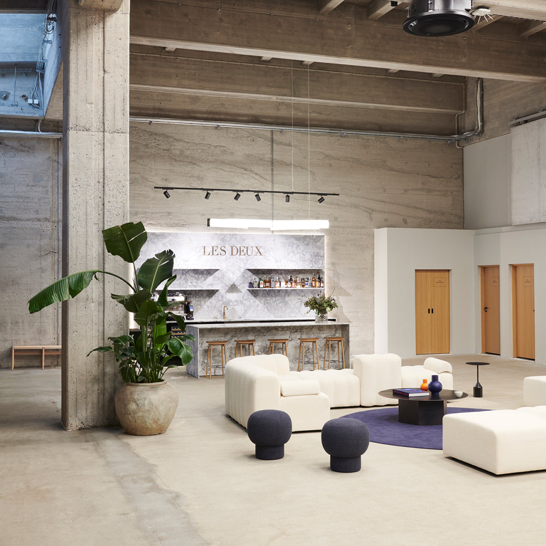 Industrial lounge with white modular seating, Les Deux marble bar, exposed concrete details, and tropical plant beside purple rug.