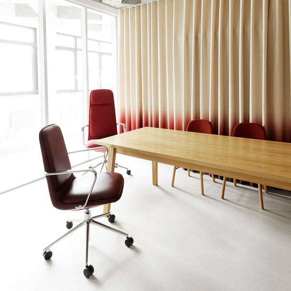 Red ergonomic chair with a sleek design, set against a light wood table and creamy gradient curtains. Minimalistic workspace decor.