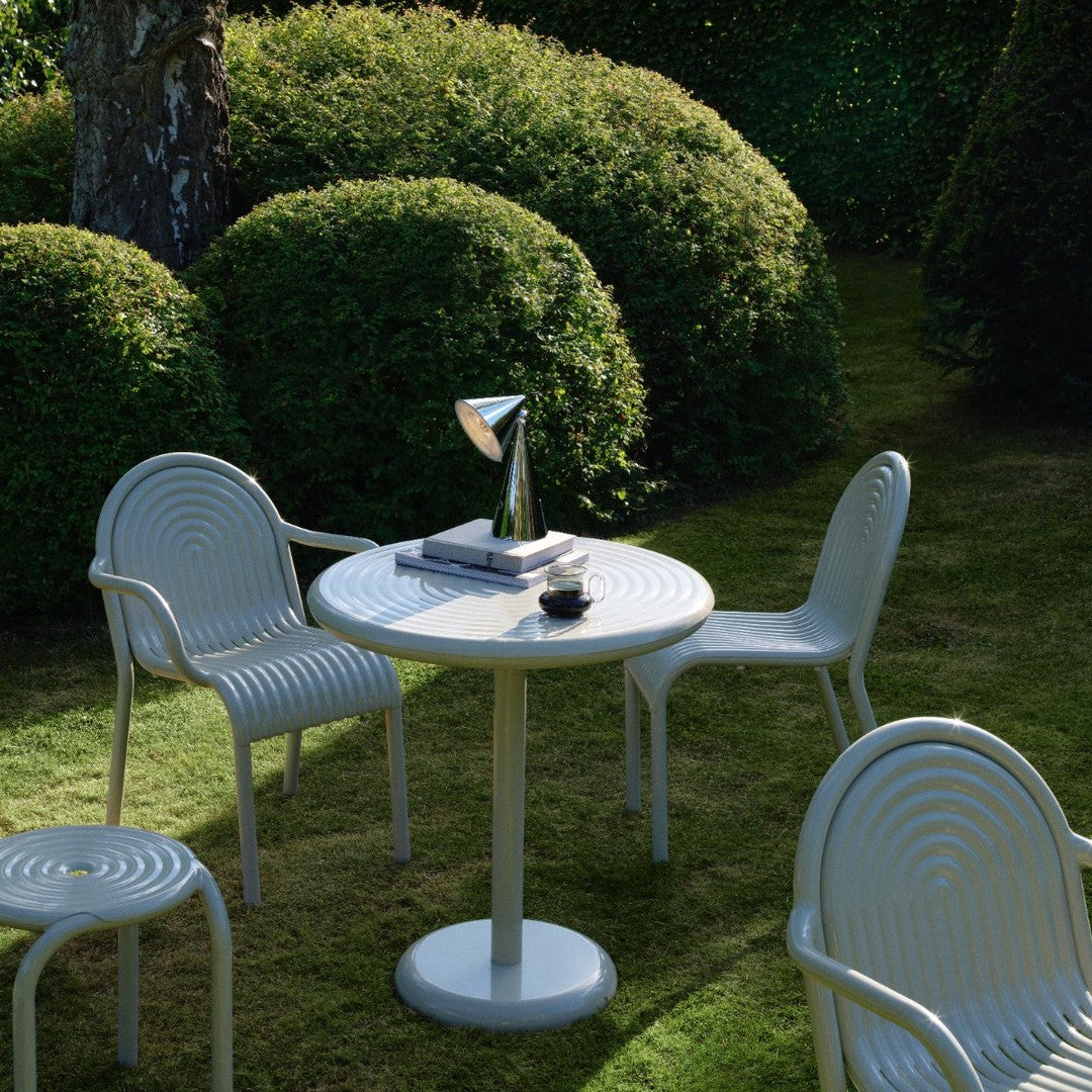 White round table surrounded by white ribbed chairs in a lush green garden, creating a serene outdoor dining space.