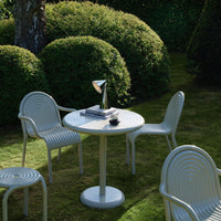 White round table surrounded by white ribbed chairs in a lush green garden, creating a serene outdoor dining space.