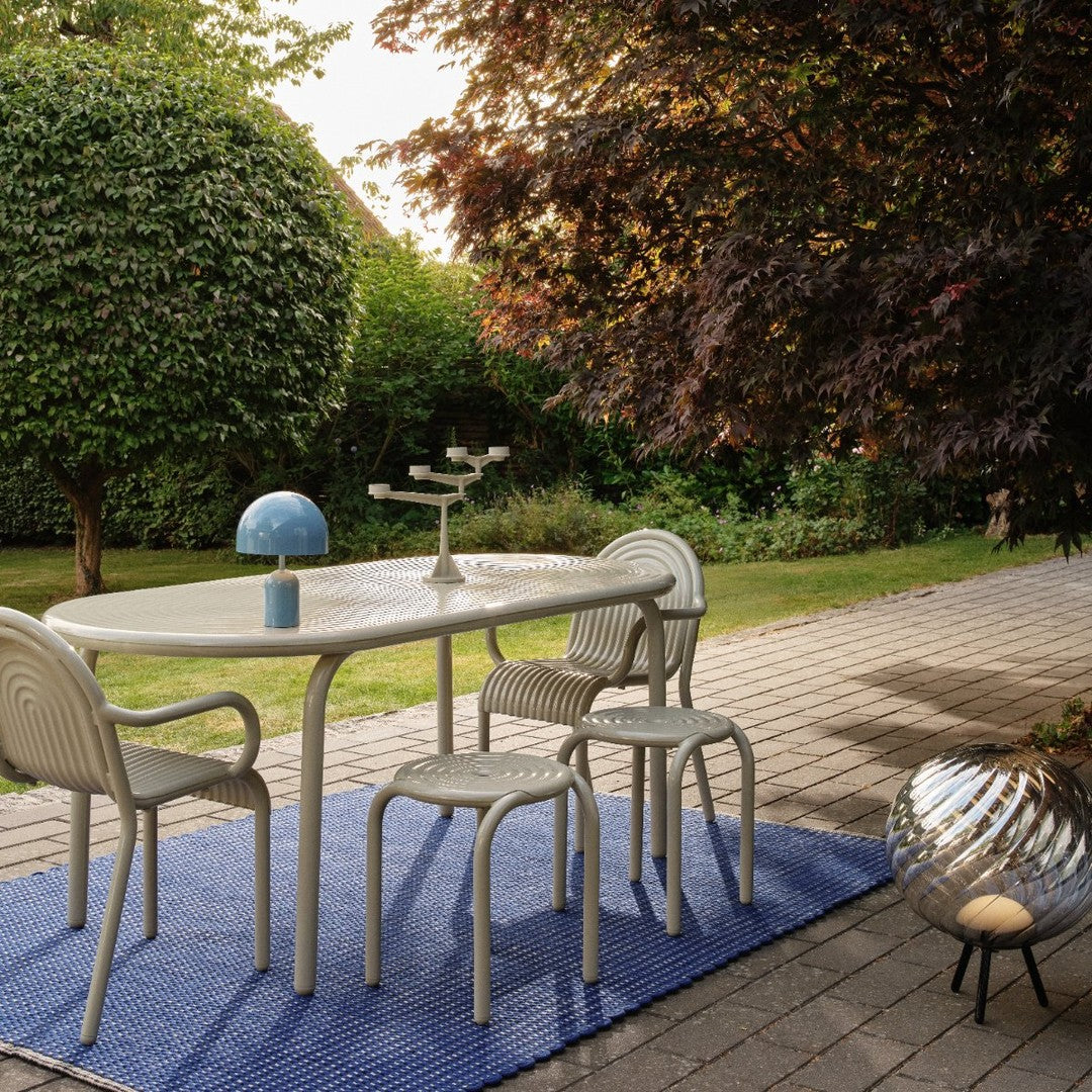 "Light gray patio table and chairs on a blue patterned rug outdoors, surrounded by green foliage and warm-toned trees."