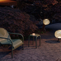 Green woven patio chair, matching table, and round lanterns with intricate patterns, set on a textured stone patio.