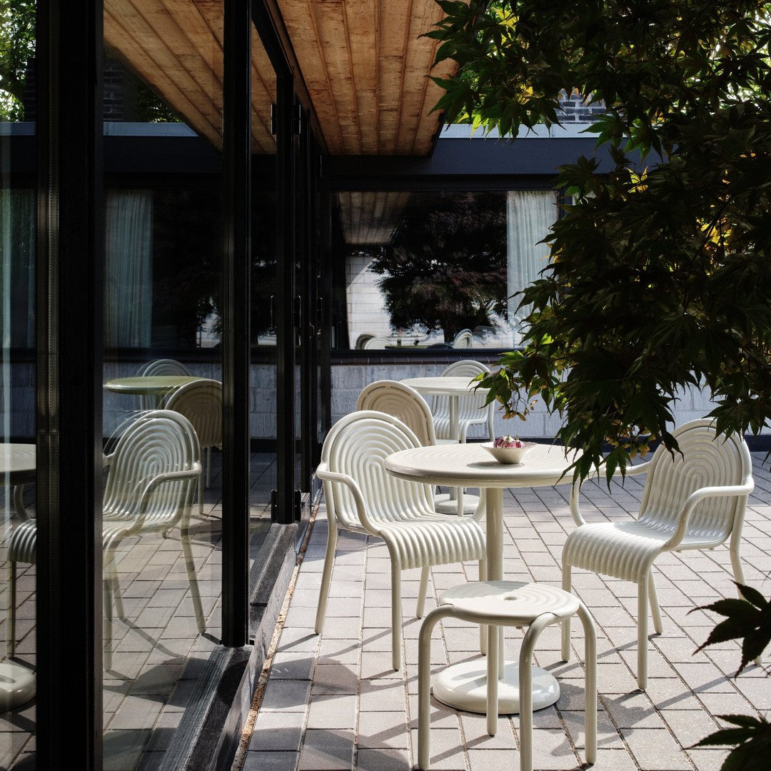 White woven chairs and a table set against a backdrop of green leaves and a reflective glass surface.