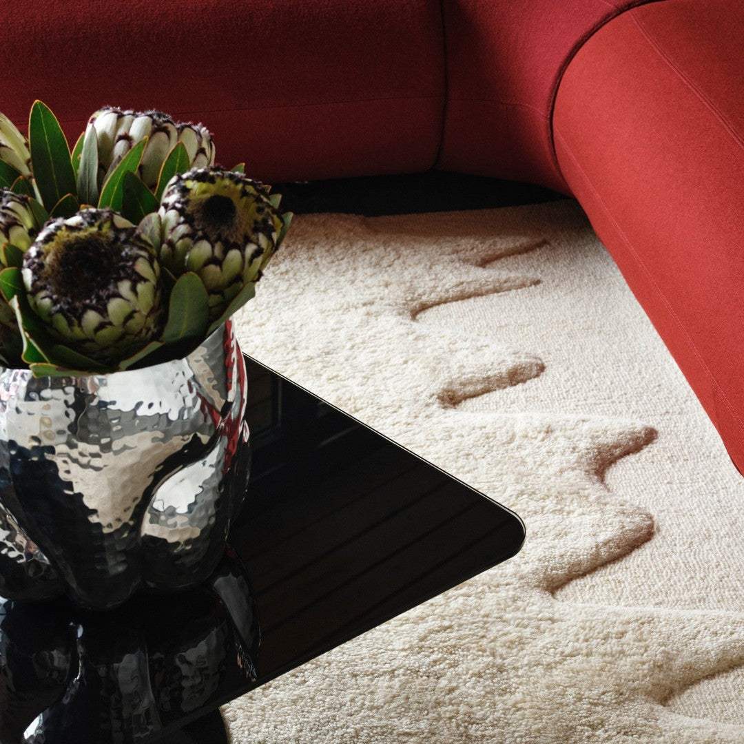 "Neutral cream rug with wave pattern, surrounded by red furniture and a metallic vase with green plants."