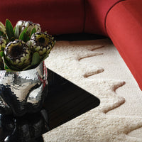 "Neutral cream rug with wave pattern, surrounded by red furniture and a metallic vase with green plants."