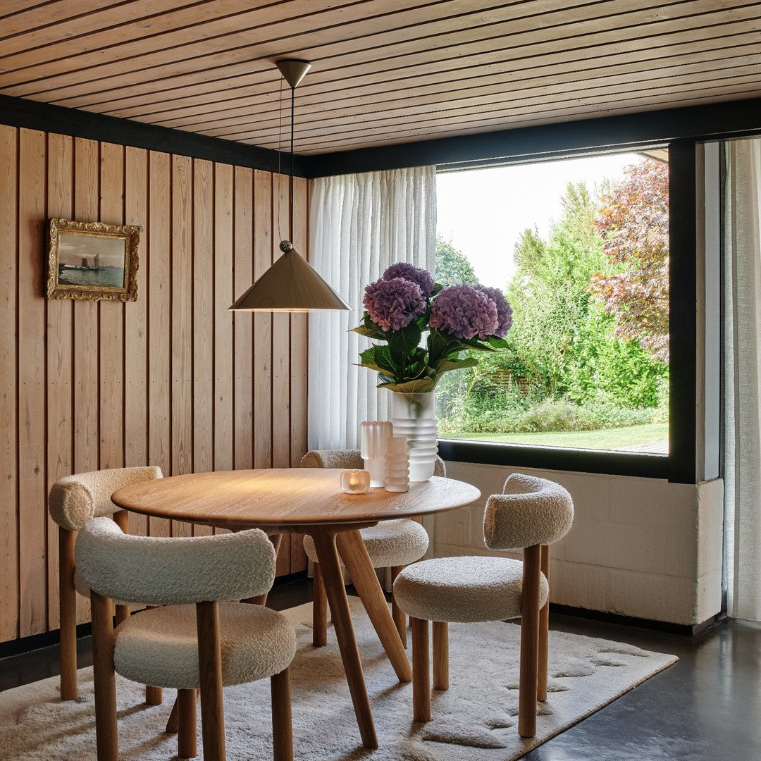 Wood-paneled walls, round wooden table, neutral-colored chairs, white curtains, and vibrant purple hydrangeas add warmth.