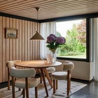 Wood-paneled walls, round wooden table, neutral-colored chairs, white curtains, and vibrant purple hydrangeas add warmth.