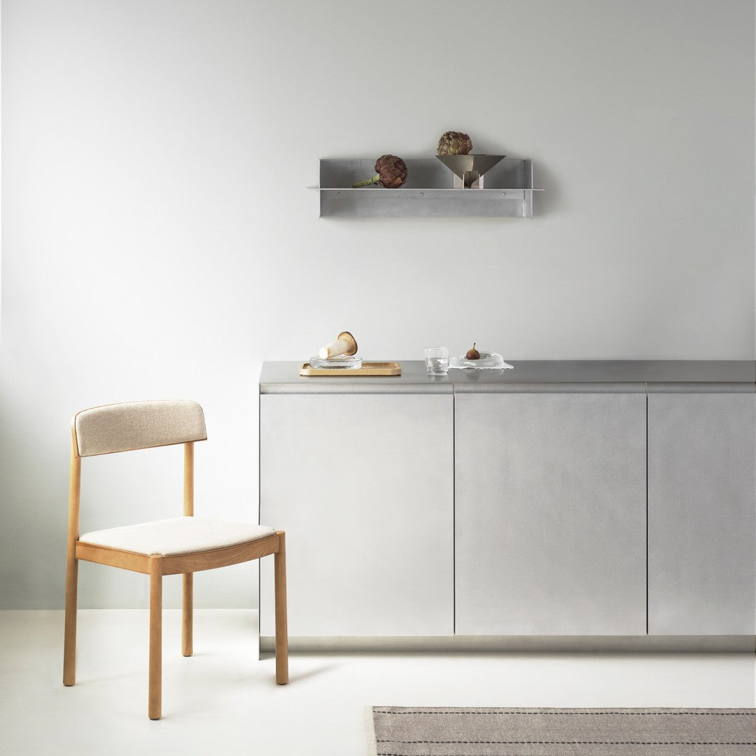 Light beige chair with wooden legs, silver cabinet, and grey wall; soft textures and minimalist design.