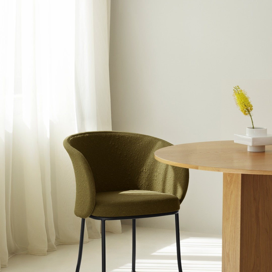 Olive green upholstered chair with a textured finish, paired with a light wooden round table. Soft, neutral backdrop.