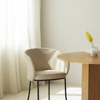 Beige textured chair with black legs beside a light wood table, paired with soft white curtains and a pale backdrop.