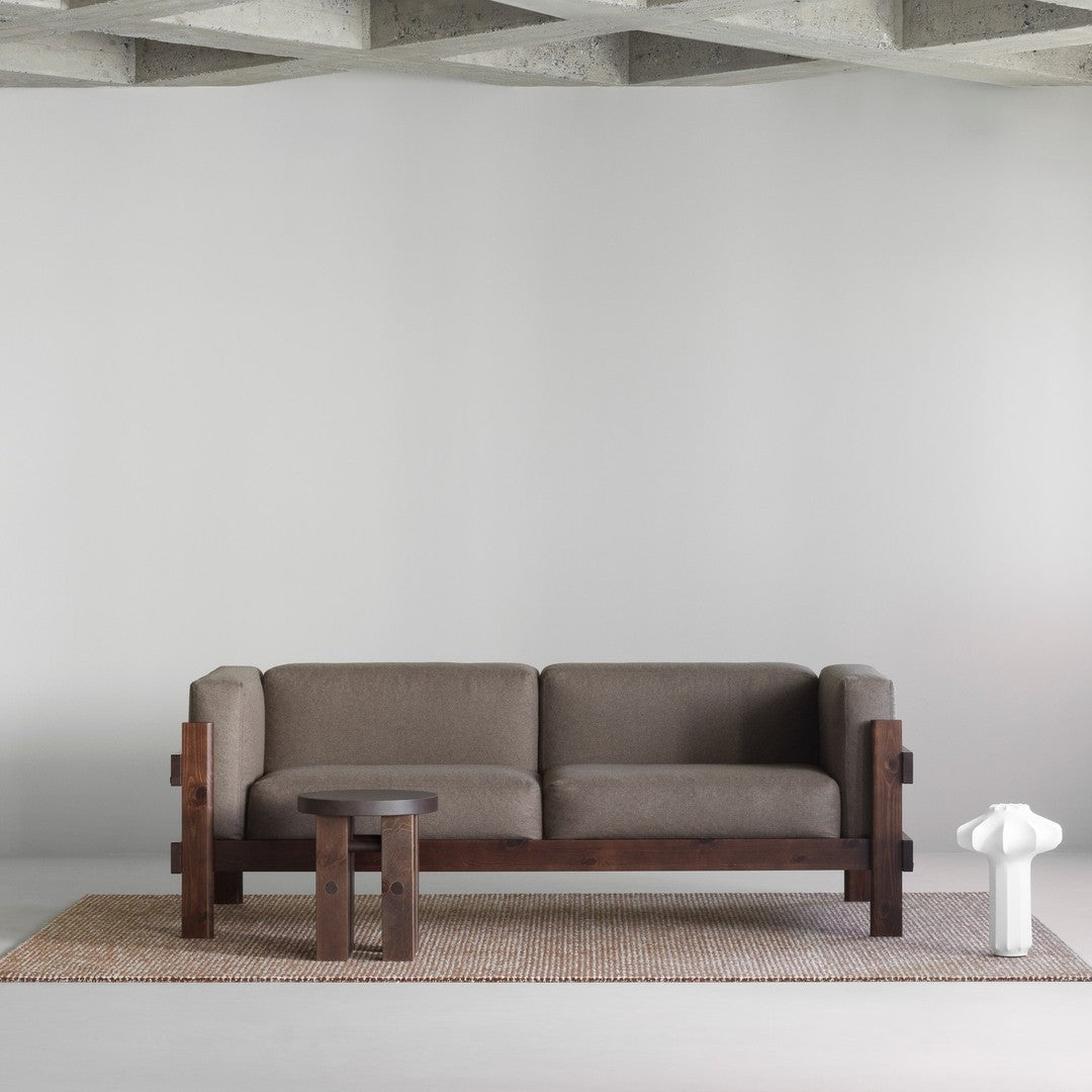 Brown fabric sofa with dark wood frame, paired with a round wooden table on a textured beige rug. Minimalist style.