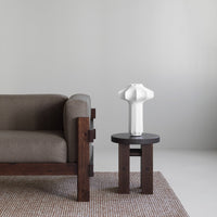 Gray couch with a white lamp on a round black table, paired with a woven beige rug. Modern minimalist design.