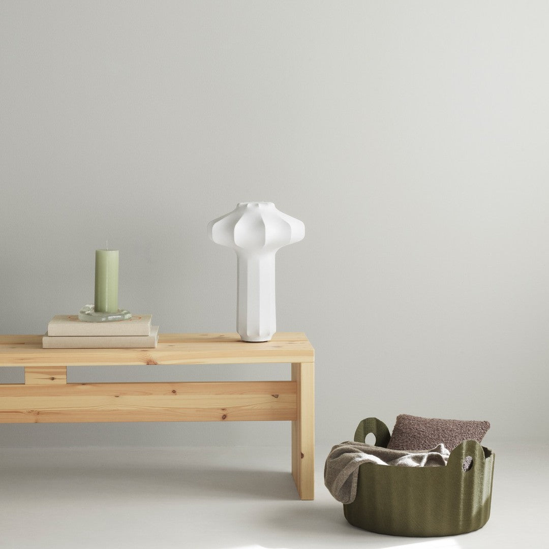 White lamp and green candle on a light wood table, with a textured olive green basket and brown cushion accent.