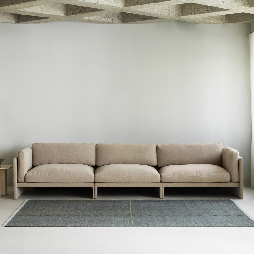Beige modern sofa with soft cushions, set on a grey patterned rug featuring a yellow accent line. Minimalist decor style.