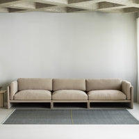 Beige modern sofa with soft cushions, set on a grey patterned rug featuring a yellow accent line. Minimalist decor style.